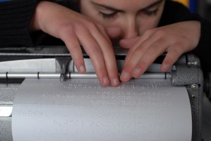 Une jeune personne est en train de lire un document en braille qui sort de sa Perkins